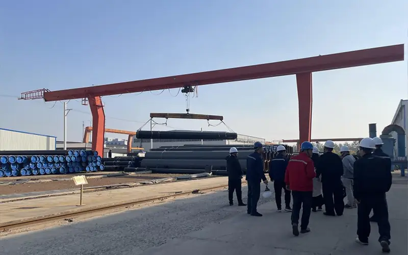 Australian client (in hard hats) audits the Allland factory yard, observing a gantry crane lifting a large coated pipe.-banner