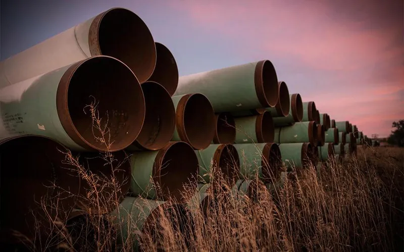 Stacked large diameter API 5L carbon steel pipes with green external coating, stored outdoors in a grassy field under a dramatic pink sunset sky.-banner