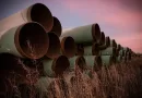 Stacked large diameter API 5L carbon steel pipes with green external coating, stored outdoors in a grassy field under a dramatic pink sunset sky.-banner