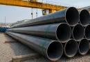 Stacks of large diameter steel pipes in an industrial yard under a gantry crane, representing API 5L PSL1 and PSL2 pipeline materials used in oil and gas infrastructure banner