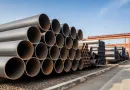 Stacks of large diameter steel pipes stored in an industrial yard illustrating ASTM A671 and ASTM A672 pipe production and supply banner