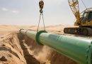 A yellow heavy-duty crane carefully lowers a green, fbe steel pipe into a trench in a vast desert landscape.